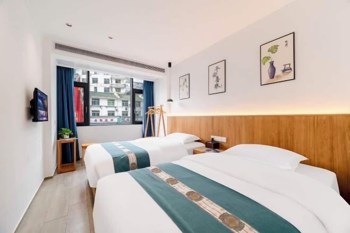 Une chambre d’hôtel avec deux lits, une télévision, une fenêtre avec des rideaux et des œuvres d’art accrochées au mur.