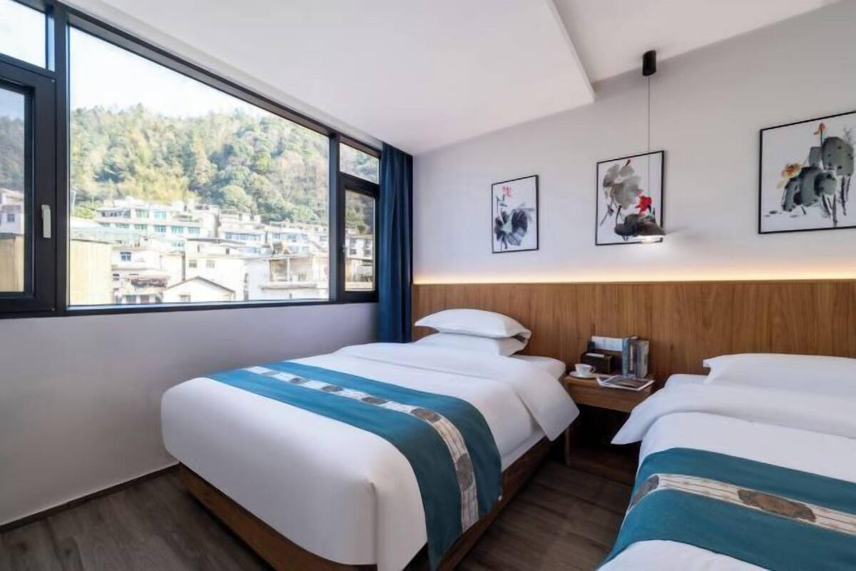 Une chambre d’hôtel avec deux lits, une grande fenêtre donnant sur des immeubles et des arbres, et des œuvres d’art accrochées aux murs.