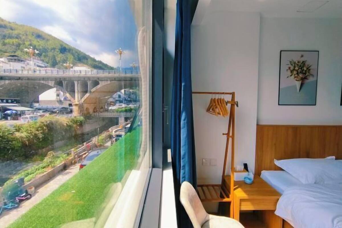Une chambre d’hôtel avec un lit, une table de chevet en bois, une chaise, une fenêtre donnant sur un pont et une route, et un tableau encadré accroché au mur.
