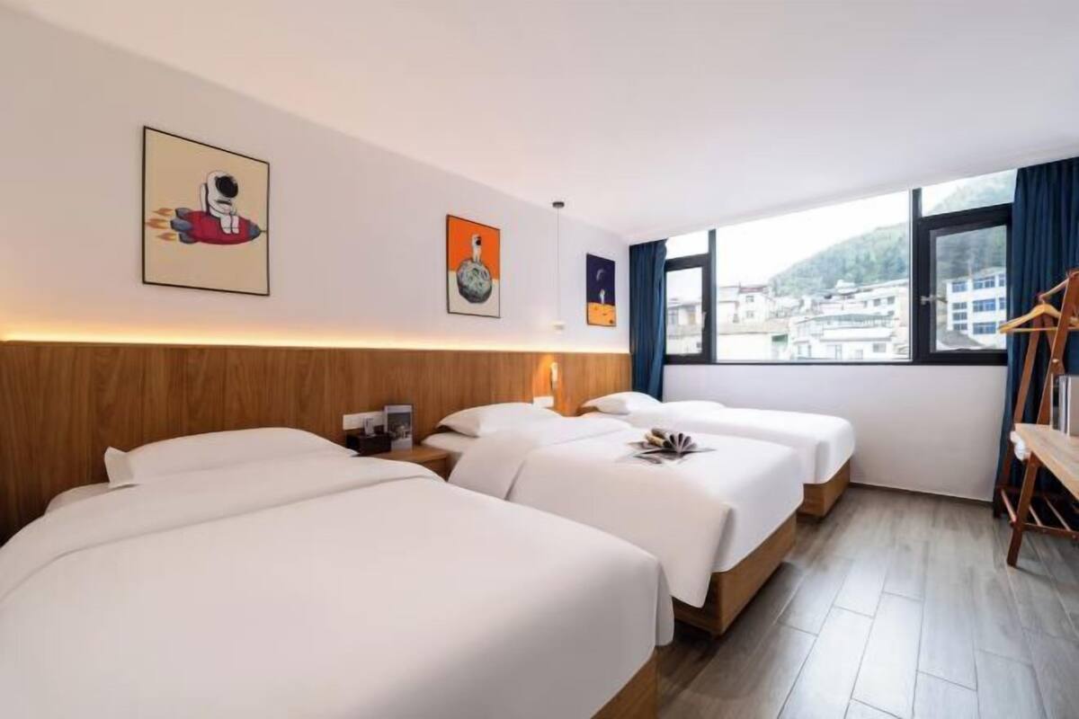 Une chambre d’hôtel avec trois lits, une tête de lit en bois, une table de chevet, une lampe et une fenêtre donnant sur des immeubles.