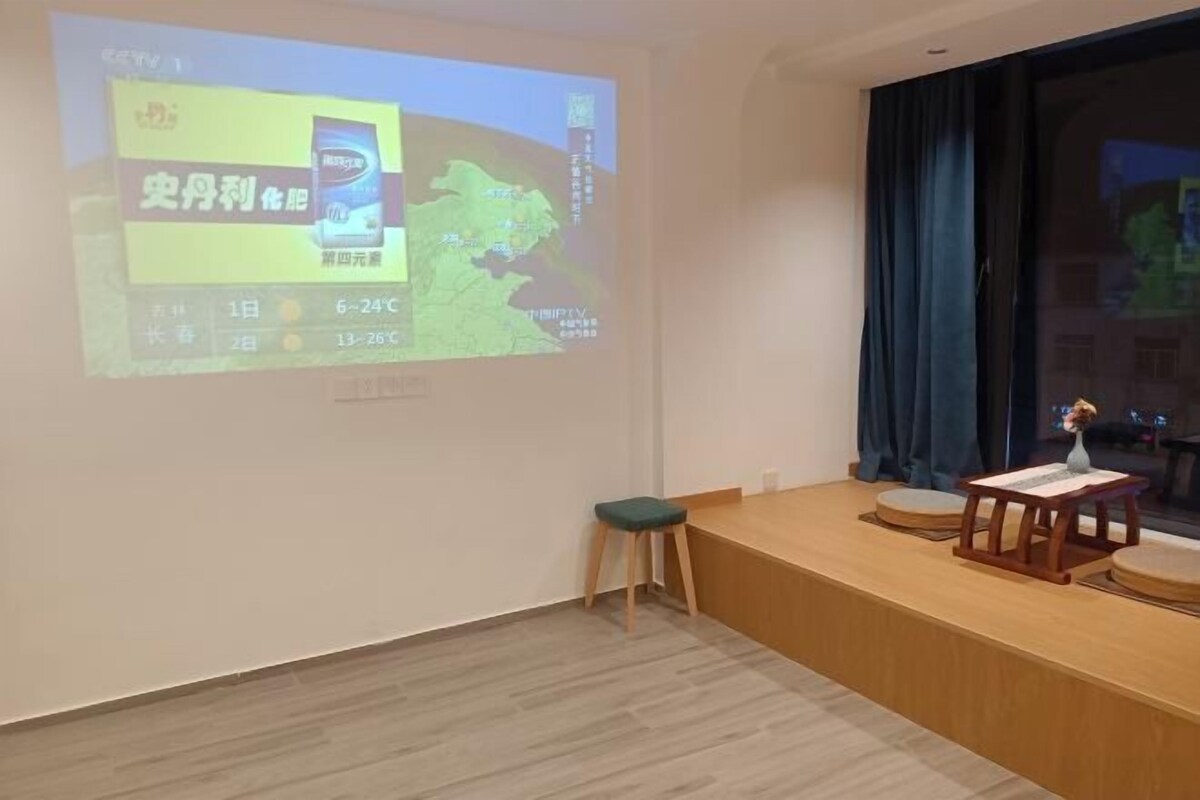 Una habitación con una pantalla de proyección que muestra un mapa y texto, un banco de madera, un taburete pequeño y una mesa con un jarrón.