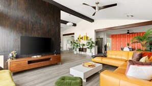 A modern living room with a large flat-screen TV, a wooden entertainment unit, a yellow sofa, green ottoman, and a white coffee table.