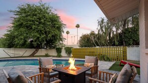 A patio with a fire pit, outdoor seating, a swimming pool, and a view of trees and a fence.