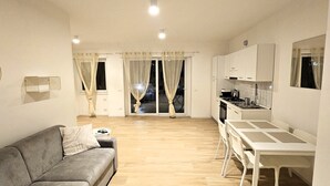 Un espace de vie compact comprenant un canapé, une table à manger, un coin cuisine et une porte-fenêtre coulissante donnant sur un balcon.