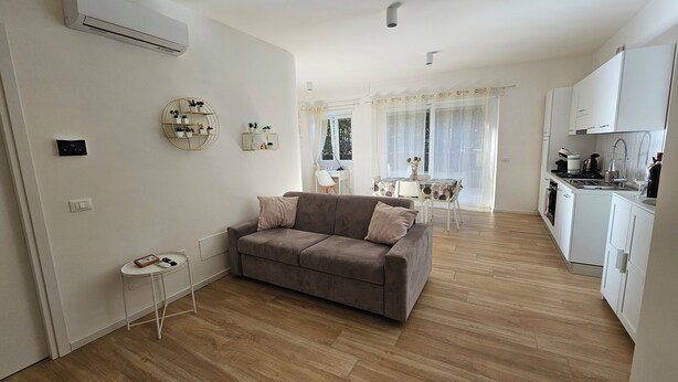 Un appartement moderne comprenant un salon, un coin repas et une cuisine. On y trouve un canapé gris, une petite table d’appoint et une table à manger blanche avec des chaises. La cuisine est équipée de meubles blancs et d’une cuisinière.