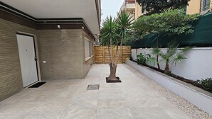 Une cour avec un sol carrelé, une porte en bois, un petit arbre et une clôture verte.