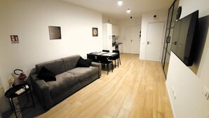 A modern living room with a grey sofa, a dining table with chairs, a small side table, and a flat-screen TV mounted on the wall. - LUX Beach Apartment BLACK (Ostia)