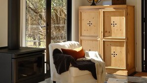 Una sala de estar acogedora con chimenea, un sillón blanco, un aparador de madera y vistas amplias a través de grandes ventanales.