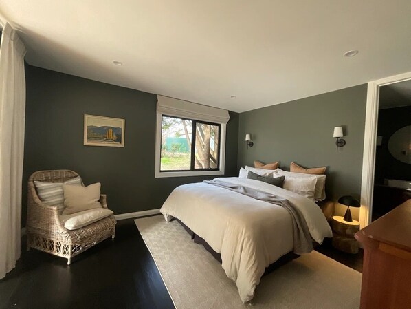 Quarto com uma cama grande, uma cadeira de vime, um quadro na parede e uma janela com vista para o verde.