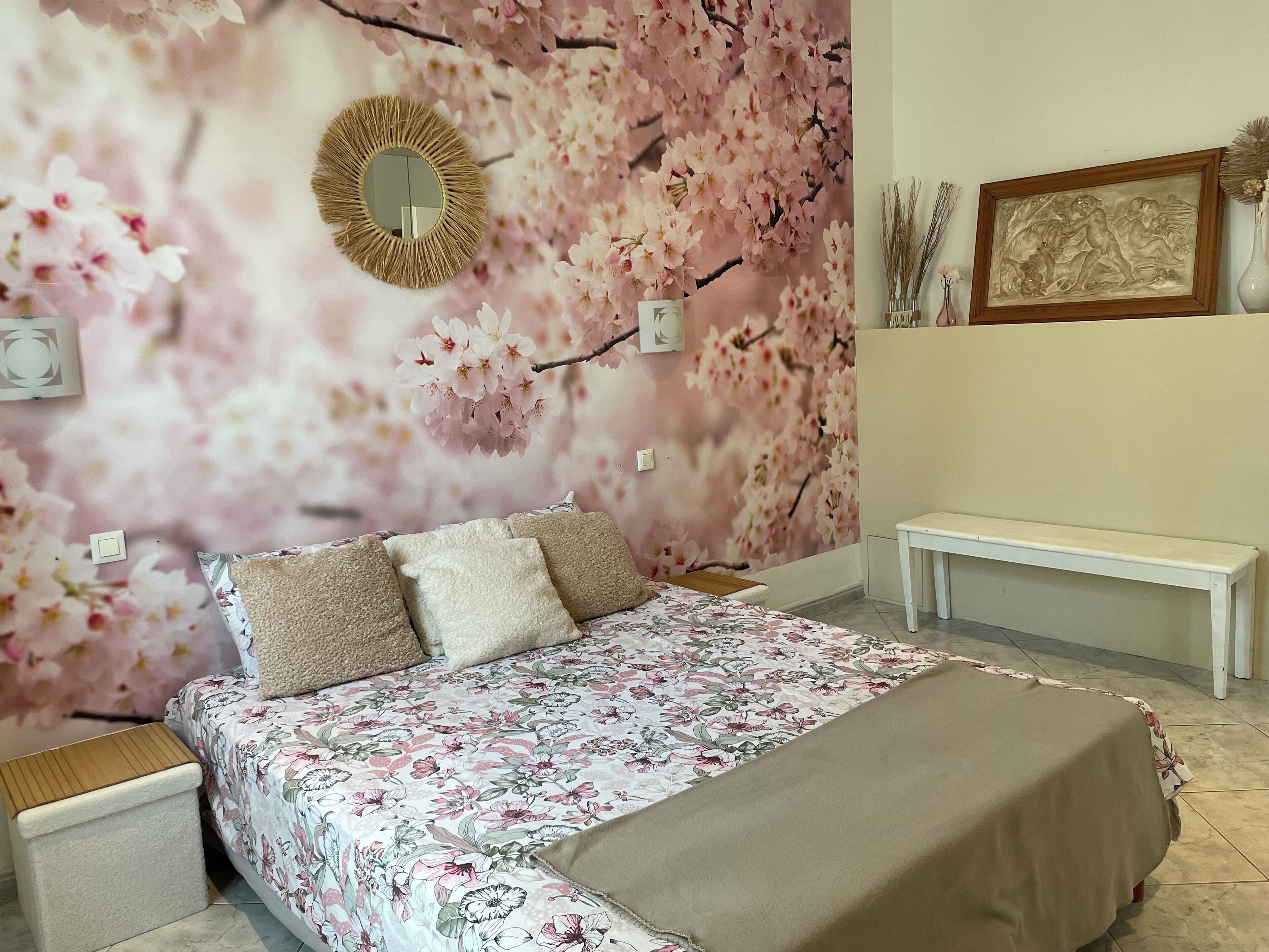 Een slaapkamer met een bloemenbedekte spreidek, een nachtkastje, een bankje en een wanddecoratie van kersenbloesems.