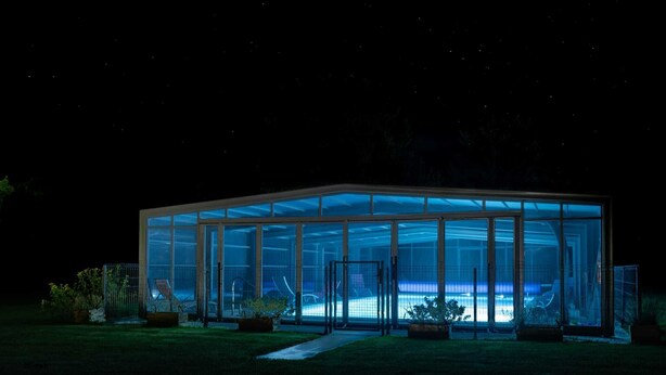 A modern glass house with a pool, surrounded by potted plants.