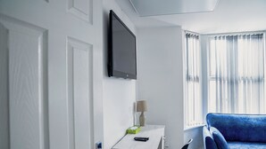 A hotel room with a flat-screen TV mounted on the wall, a small desk with a lamp, a blue sofa, and a window with sheer curtains. - 2Bed Apartment Near City Centre (Sleeps 4, with free parking) (Coventry)