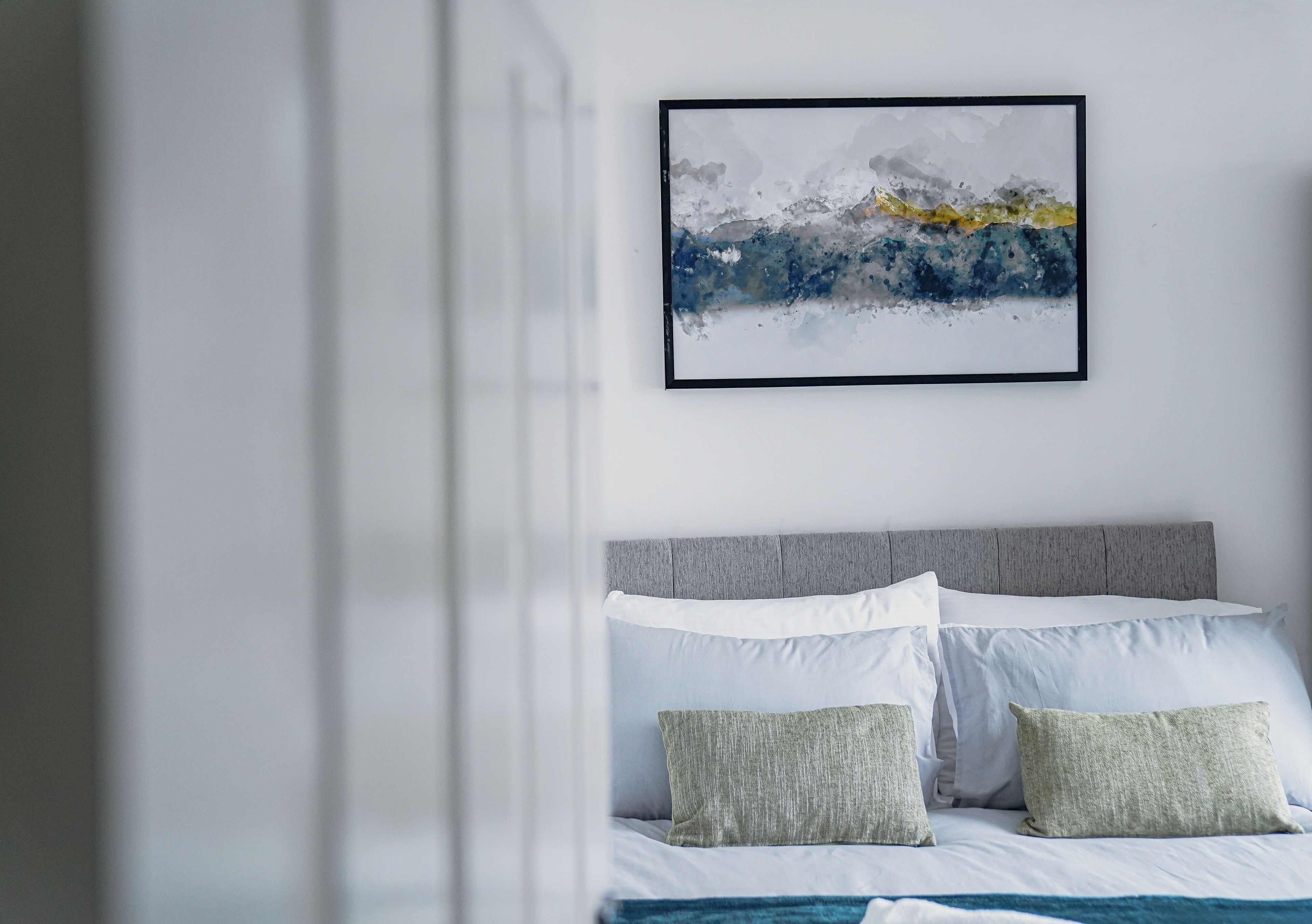 A modern bedroom with a grey headboard, white bedding, and a framed abstract painting on the wall.