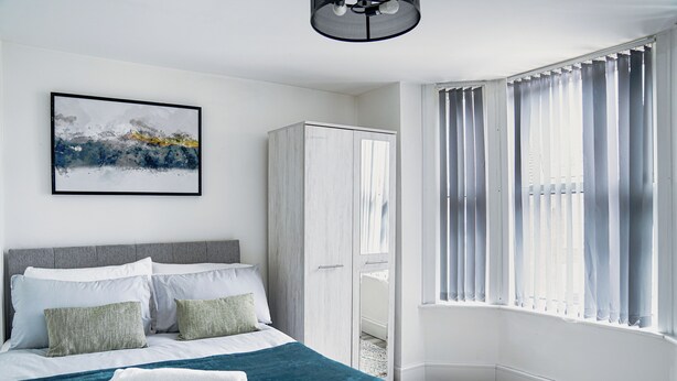 A bedroom with a bed, a wardrobe, a window with curtains, and a painting on the wall. - 2Bed Apartment Near City Centre (Sleeps 4, with free parking) (Coventry)