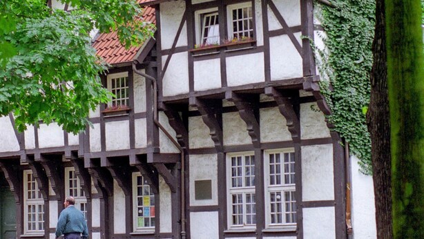 Ein traditionelles Fachwerkhäuschen mit Ziegeldach, eine Kopfsteinpflasterstraße und eine Person, die davor geht.