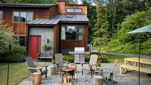 Ett modernt hus med en träfasad, en röd dörr och en uteplats med utemöbler och en grill.