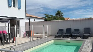 Ein Schwimmbad mit Holzterrasse, weißem Zaun und einem Gebäude mit blauen Fensterläden.