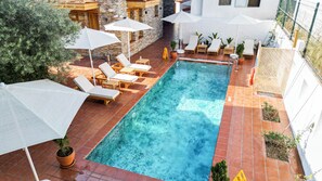 A rectangular swimming pool with a tiled edge, surrounded by lounge chairs and umbrellas, set against a stone wall and a white building.