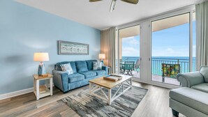 A spacious living room with a blue sofa, a coffee table, and a view of the ocean through large windows. - 🌊Beachfront with Gorgeous Views-Aqua Resort 905🌊 (Panama City Beach)