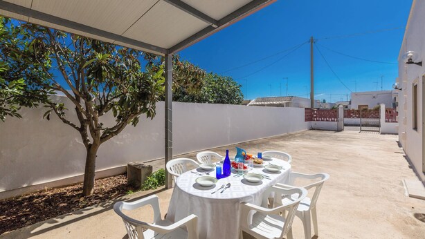 Een terras buiten met een ronde eettafel, omringd door witte plastic stoelen.