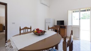Een eetgedeelte met een ronde tafel, stoelen, een witte tafelkleed, een fruitmand, een airconditioner, een televisie en een raam met uitzicht.