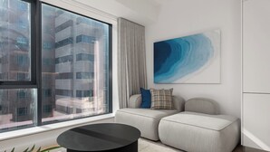 A modern living room with a large window, a round black coffee table, a grey sofa with a blue pillow, and a blue abstract painting on the wall. - The Rubik | Designer One-Bedroom in Cape Town (Cape Town)