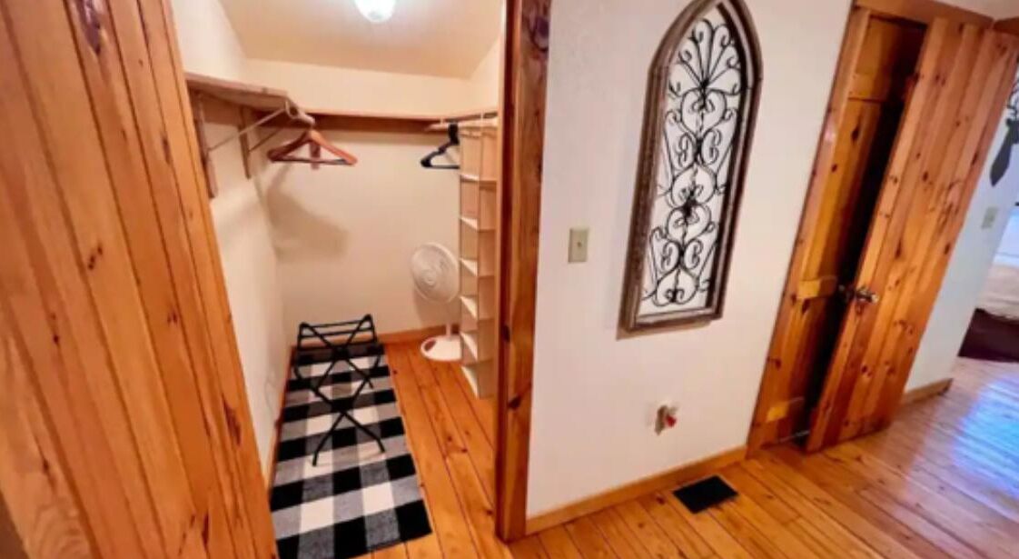 Un corridoio con pavimento in legno, un tappeto a scacchi, un appendiabiti, una ventola e un telaio di ferro decorativo alla finestra.
