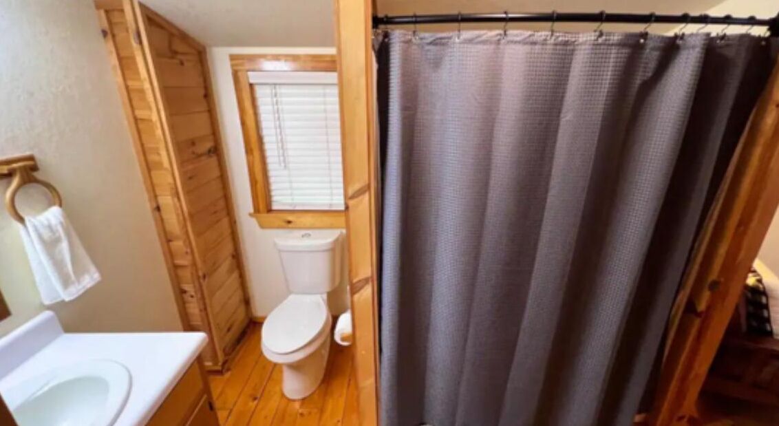 Ein Badezimmer mit Holzboden, Toilette in Weiß, Holztür und Duschvorhang.