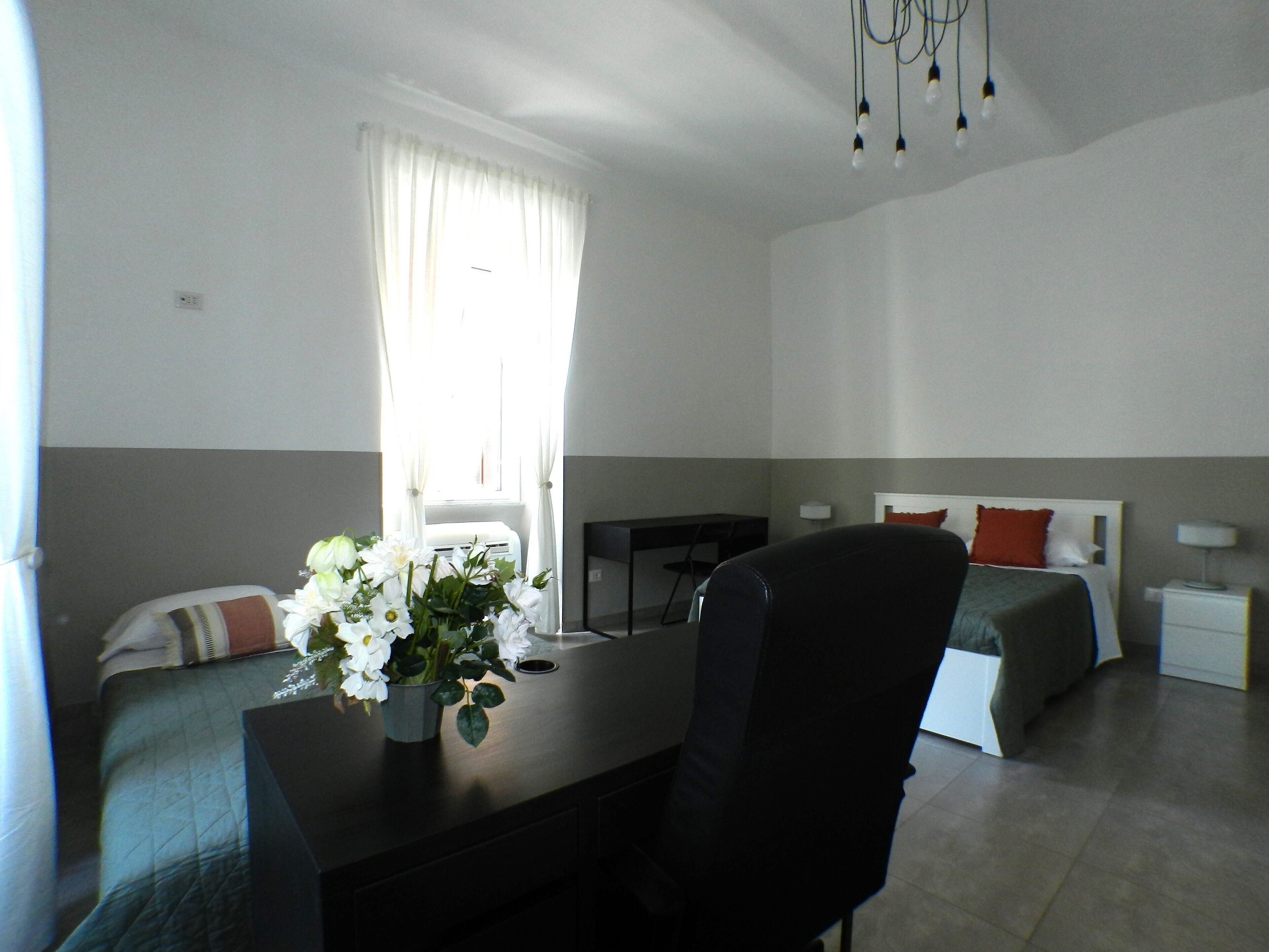 A hotel room with a desk, two beds, a chair, a desk lamp, and a window with curtains.