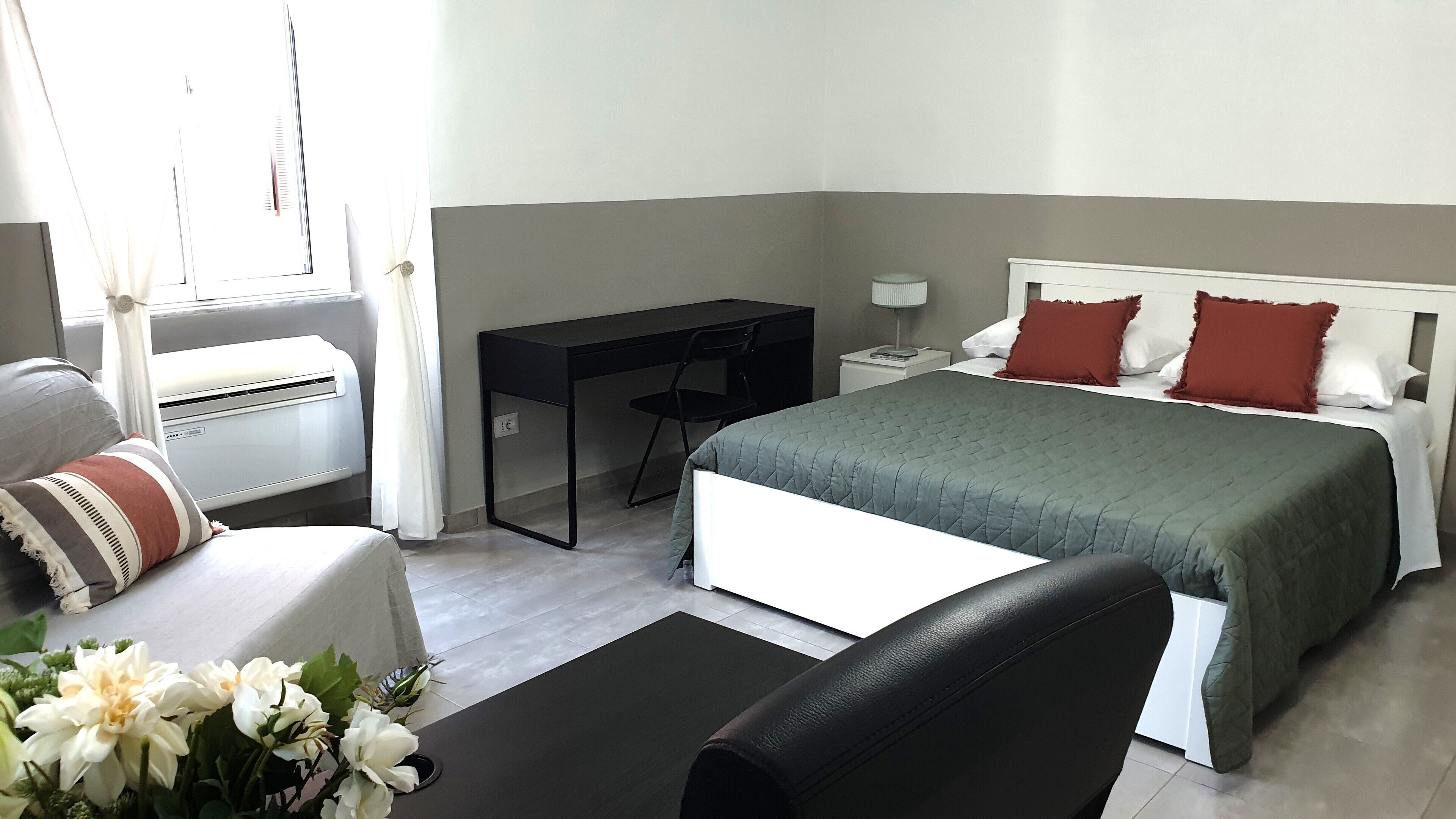 Een hotelkamer met een bed, een bureau, een stoel, een klein tafeltje, een nachtkastje, een lamp en een raam met gordijnen.
