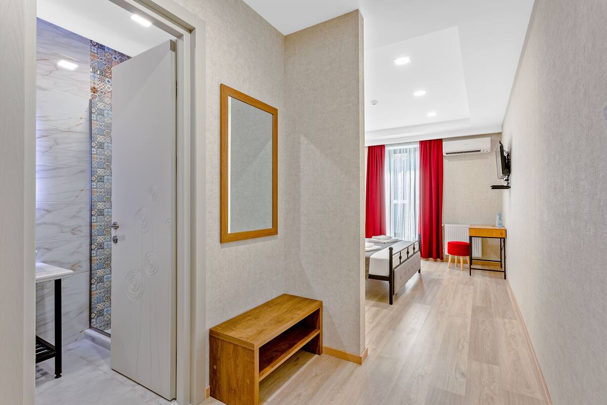 Ein modernes Hotelzimmer mit einer hölzernen Bank, einem Spiegel, einem kleinen Tisch und einem Bett mit rotem Vorhang.