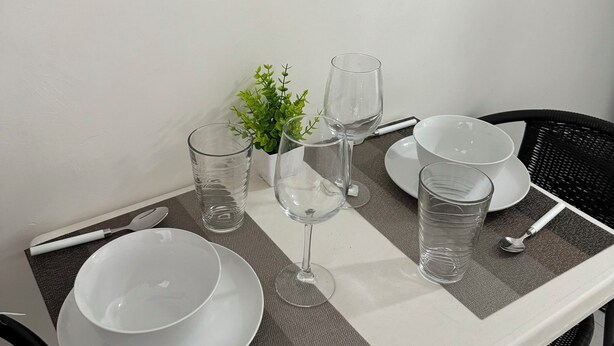 Une table à manger dressée avec de l’assiette blanche, des bols, des verres et des couverts, agrémentée d’une petite plante en pot.