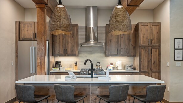 Una cocina moderna con armarios de madera, una isla central con taburetes altos, electrodomésticos de acero inoxidable y lámparas colgantes.