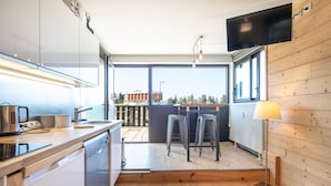 Eine moderne Küche mit Spülmaschine, Kaffeemaschine und an der Wand montiertem Fernseher. Es gibt Hocker und ein Fenster mit Blick auf ein Gebäude.