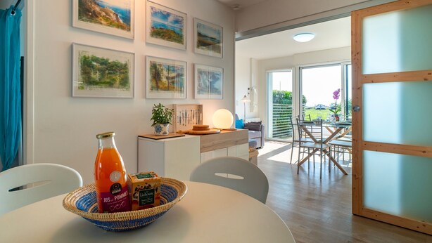 A dining area with a table, chairs, and a basket with a bottle and a box. There are framed pictures on the wall and a door leading to an outdoor area.