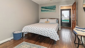 A bedroom with a bed, a ceiling fan, a painting, a door, and a view of the outside.