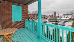 A turquoise balcony with a wooden chair, brick wall, and a view of a town with a lighthouse.