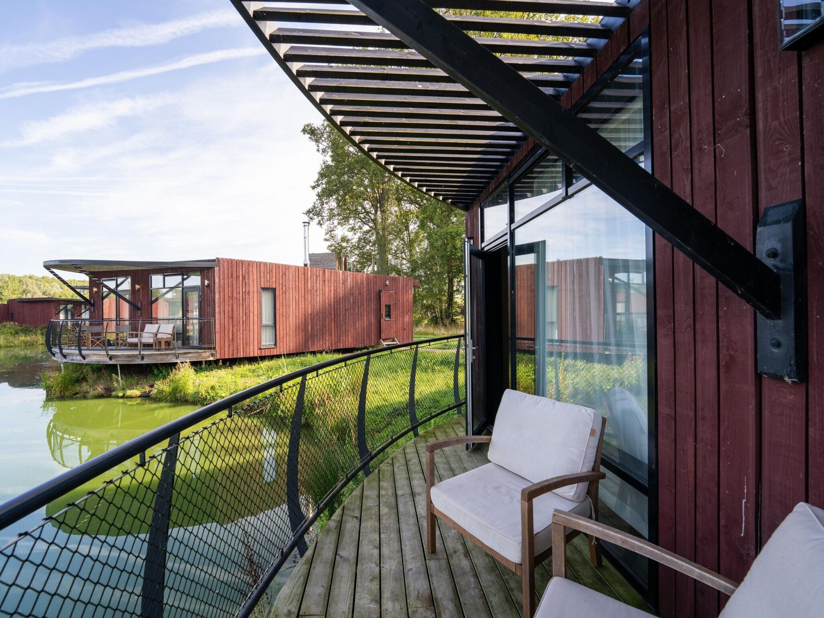 A wooden deck with a glass railing overlooking a body of water, a wooden deck chair, and a modern building with large windows.