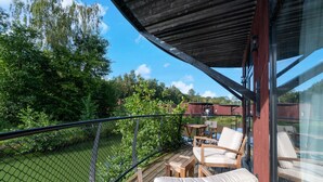 Une terrasse en bois avec des meubles d’extérieur, une véranda vitrée et une vue sur une rivière et des arbres.