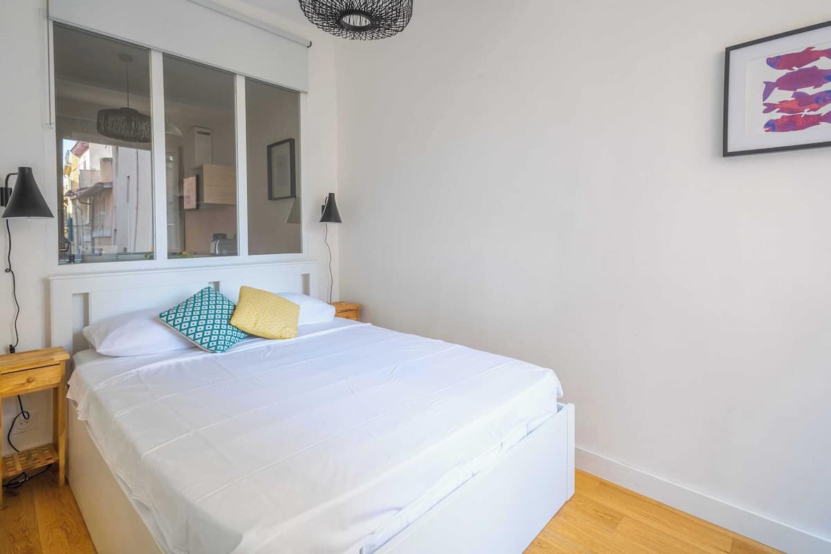 Une chambre avec un grand lit, une fenêtre, deux tables de chevet et une lampe fixée au mur.