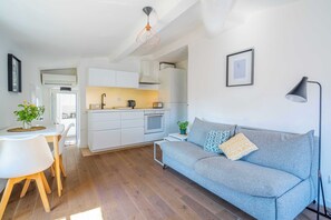 Une cuisine moderne avec des meubles blancs, un coin repas avec une table et des chaises blanches, et un canapé gris agrémenté d’une plante et d’une lampe.