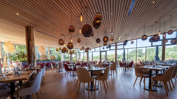 A spacious restaurant with wooden floors and a high ceiling, featuring round hanging lights and large windows offering a view of the outdoors. - Charming Forest Cottage (Antoing)