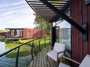 A wooden deck with a glass railing overlooking a body of water, a wooden deck chair, and a modern building with large windows. - Relaxing Bungalow Stay (Antoing)