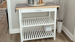 Una consola de madera blanca con una maceta de metal y portavelas, un estante blanco en la parte inferior y dos cuadros de mariposas en la pared.