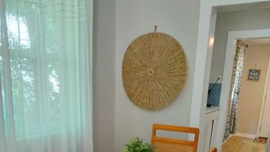 Un comedor con una mesa de madera, sillas blancas, un tapiz tejido en la pared y una planta pequeña sobre la mesa.