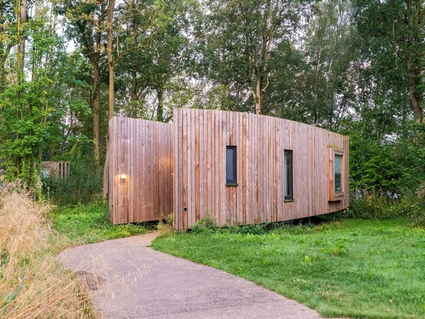 Una baita di legno con due finestre e un piccolo portico, immersa tra alberi e raggiungibile tramite un sentiero sterrato.