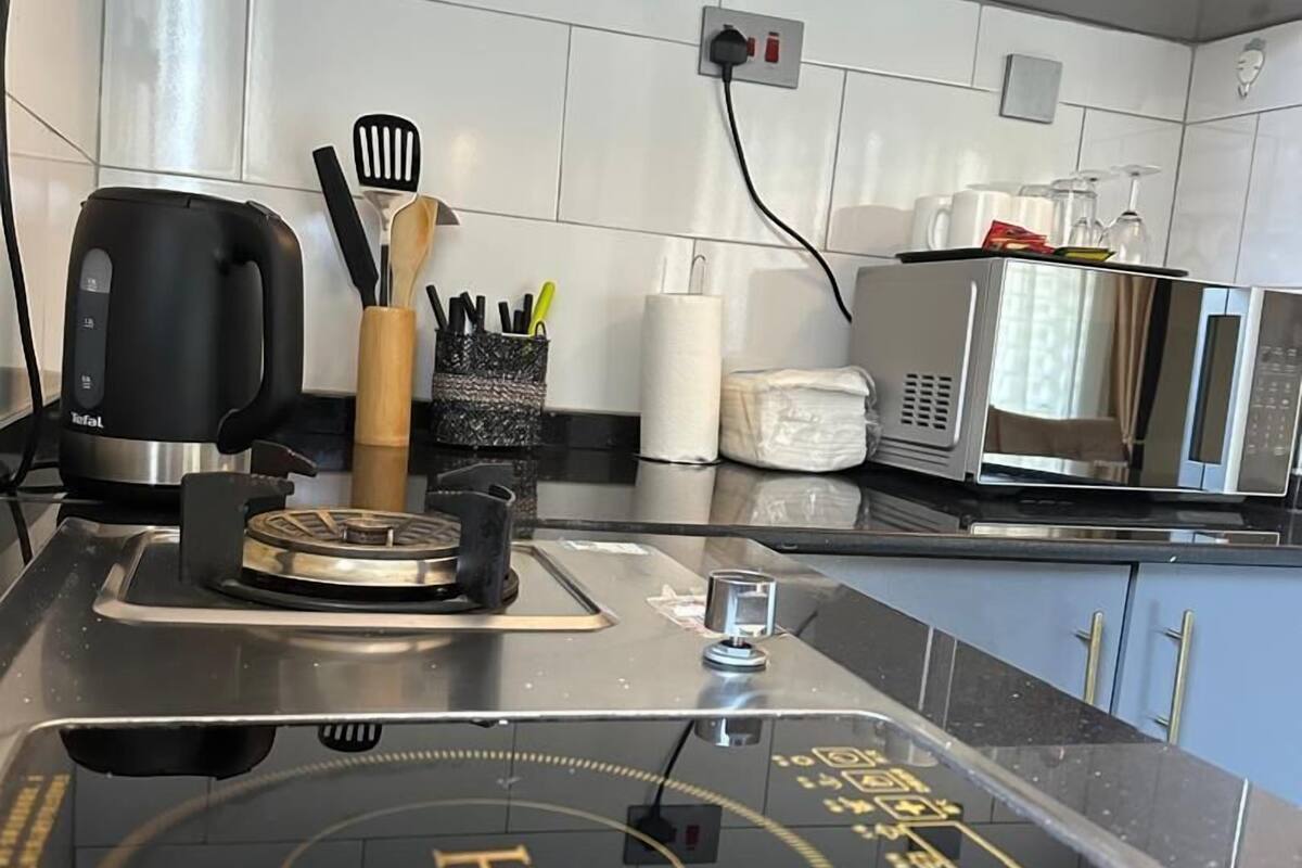 A kitchen with a black induction cooktop, a kettle, a microwave, and a coffee maker. There are utensils in a holder and a roll of paper towels on the counter.