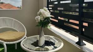 Ein Balkon mit einem weißen Korbstuhl, einem kleinen runden Tisch mit einer Blumenvase und einer schwarzen Trennwand.