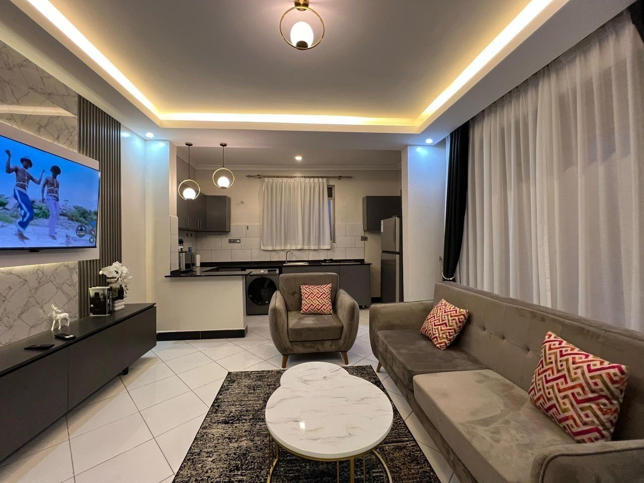 Ein modernes Wohnzimmer mit einer Couch, einem Couchtisch und einem Fernseher. Im Hintergrund ist eine Küche zu sehen.