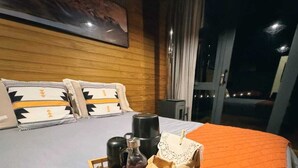 Habitación de hotel con una cama, cabecera de madera, mesita de noche, ventana con cortinas y una cesta de frutas sobre la cama.